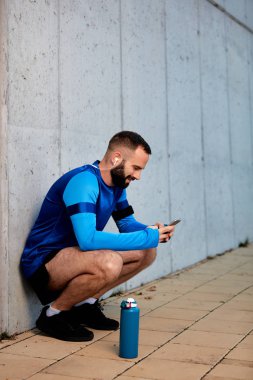 Genç adam çömelmiş ve akıllı telefon kullanarak sakallı. Onun yanında şişe su ile. Arka duvarda. Sağlıklı yaşam konsepti.