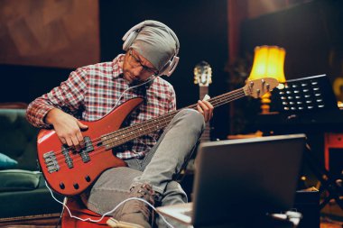 Kap ve bas gitar çalmaya kulaklık olan adam. Ftont onu içinde laptop. Ev stüdyosu iç.