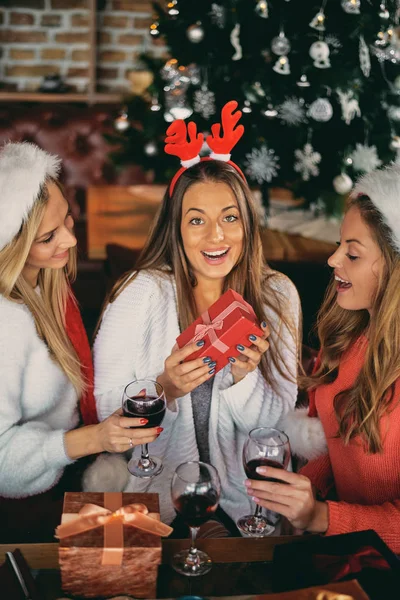 Friends giving gifts to each other and drinking wine while sitting at table. In background Christmas tree. Christmas holidays concept.