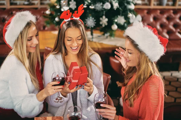 Friends giving gifts to each other and drinking wine while sitting at table. In background Christmas tree. Christmas holidays concept.