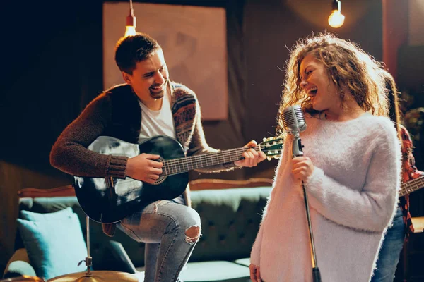 Band practice for the show. Woman with curly hair holding microphone and singing while man in background playing acoustic guitar. Home studio interior.