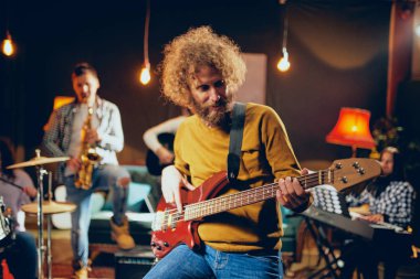Bas gitarist sandalyede otururken gitar çalmak kapatın. Arka plan sanatçısı ve saksafon piyano Player'da. Ev stüdyosu iç.