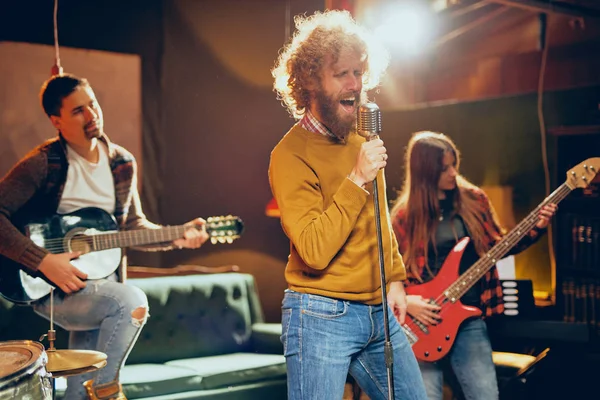 Band practicing for the gig.  Male singer with curly hair holding microphone and singing. In background band playing instruments. Home studio interior.