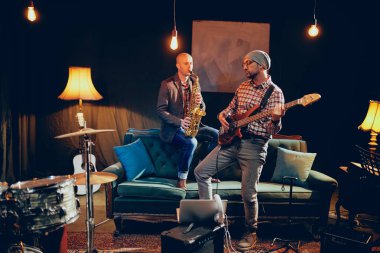 Ev stüdyosunda dururken bas gitar çalmaya kafasına şapka ile genç beyaz adam. Arka plan saksofoncu onun saz.