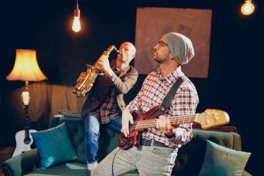 Ev stüdyosunda dururken bas gitar çalmaya kafasına şapka ile genç beyaz adam. Arka plan saksofoncu onun saz.