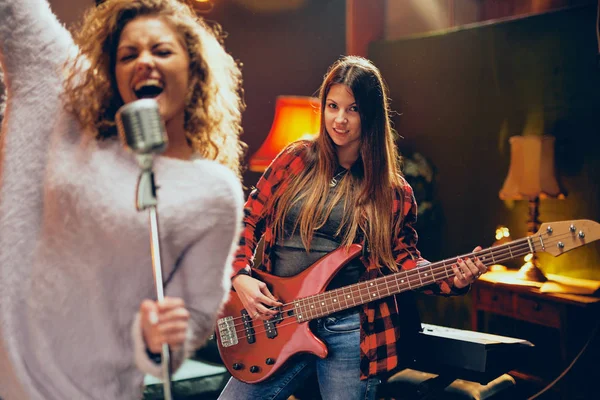 Two women practicing for the gig. Woman with curly hair holding microphone and singing while other one playing bass guitar. Home studio interior.