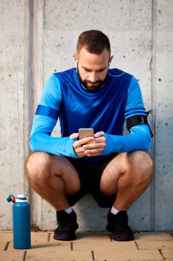 Genç adam çömelmiş ve akıllı telefon kullanarak sakallı. Onun yanında şişe su ile. Arka duvarda. Sağlıklı yaşam konsepti.