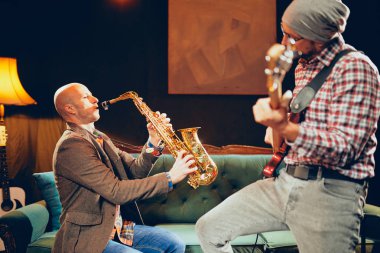 Ev stüdyosunda otururken bas gitar çalmaya kafasına şapka ile genç beyaz adam. Onun yanında onun saz sanatçısı.