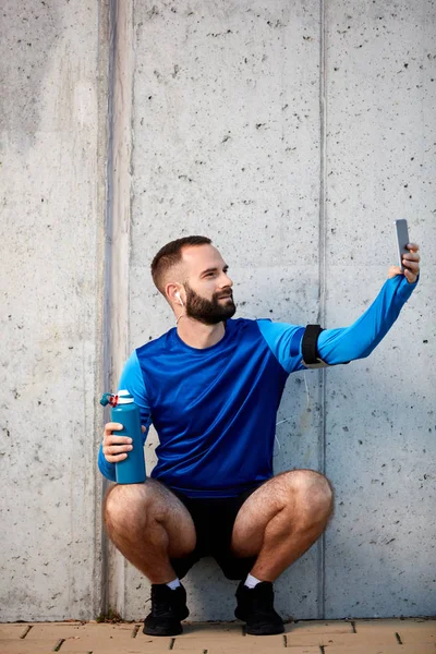 Hombre Barbudo Joven Agachándose Usando Teléfono Inteligente Sosteniendo Botella Con — Foto de Stock