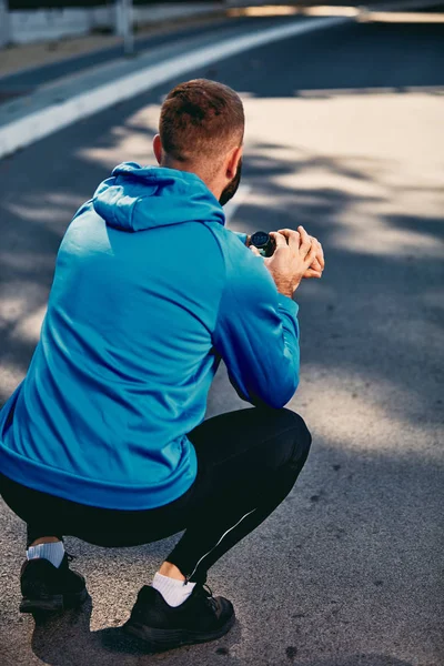 Hombre barbudo arrodillado en la calle y comprobando el latido del corazón en un reloj inteligente. Auriculares en los oídos. concepto de estilo de vida saludable . — Foto de Stock