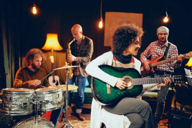 Şarkı ve bağdaş ile sandalyede otururken gitar çalmak karışık ırk kadın. Arka plan davulcu, saksofoncu ve bas gitarist.