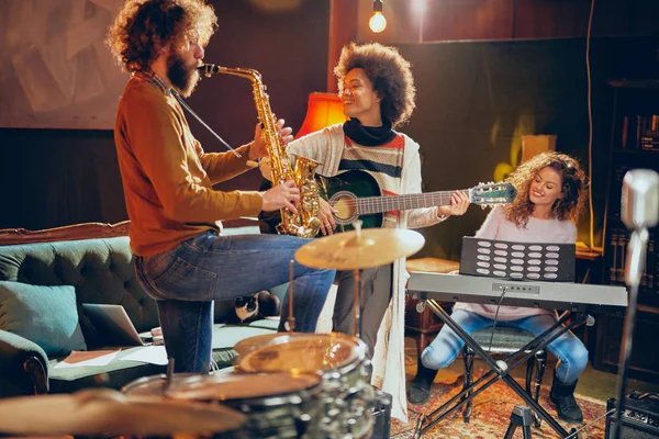 Mixed race woman playing acoustic guitar while man playing saxophone. Home studio interior. In background woman playing clavier.