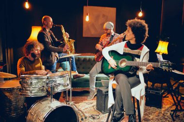 Akustik gitar çalmak ve ev stüdyosunda bağdaş ile oturan karışık ırk kadın. Grubun geri kalanı davul, saksafon ve bas gitar artalanda çalışır durumda.