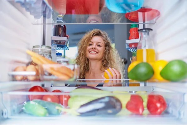 Женщина, стоящая перед открытым холодильником и принимающая апельсиновые фрукты — стоковое фото