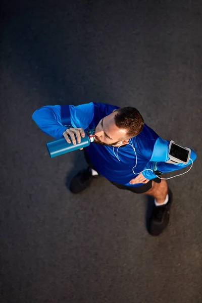 Vista superior de un joven corredor barbudo parado en la calle en ropa deportiva y agua potable. Alrededor del brazo caja del teléfono inteligente . — Foto de Stock