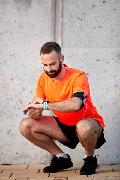 Sporty young man crouching and setting timer on wristwatch. Healthy lifestyle concept.