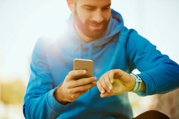 Primer plano del joven barbudo mirando el reloj inteligente y sosteniendo el teléfono inteligente en otra mano . — Foto de Stock