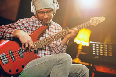 Beyaz delikanlı ev stüdyosunda otururken bas gitar çalmaya kafasına şapka ile.