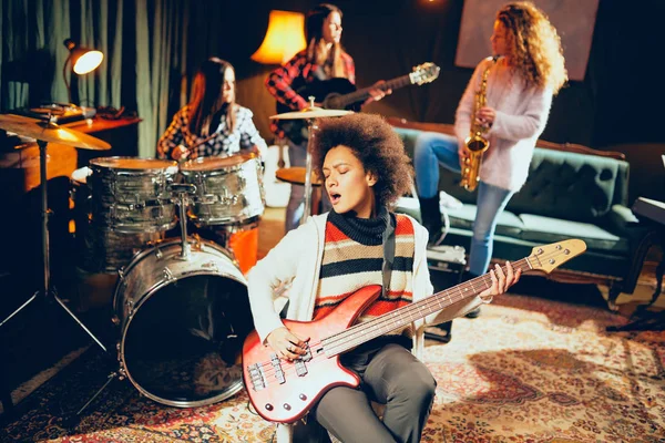 Girls playing jazz music. In foreground one woman playing bass guitar and in background other three playing acoustic guitar, saxophone and drums. Home studio interior.