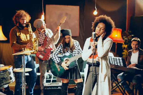 Donna di razza mista che canta. In banda di sottofondo a suonare strumenti. Home studio interno . — Foto Stock