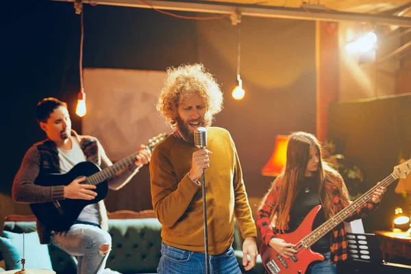 Kapela cvičí na koncert. Zpěváka s kudrnatými vlasy hospodářství mikrofon a zpěvu. V pozadí kapela hrající nástroje. Domácí studio interiér. — Stock fotografie