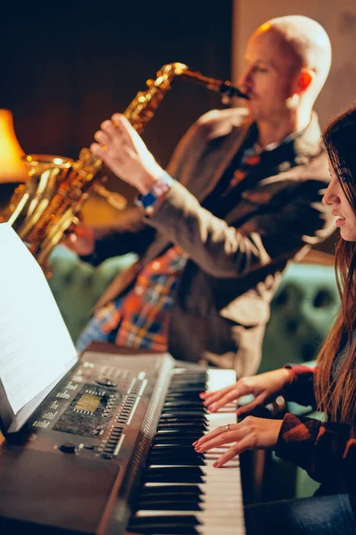 Fiatal kaukázusi nő, hosszú barna haj játszik elektromos zongora és háttér szaxofonos játszik ő eszköze. Belső Home studio. — Stock Fotó