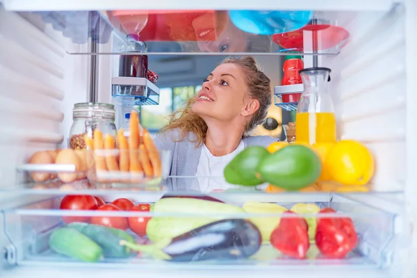 Donna in piedi davanti al frigo pieno di generi alimentari e in cerca di qualcosa da mangiare. Foto scattata dall'interno del frigorifero . — Foto Stock