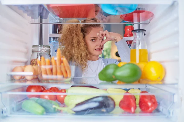 Donna in piedi di fronte al frigo aperto e tenendo il naso a causa del cattivo odore. Foto scattata dall'interno del frigo pieno di generi alimentari . — Foto Stock