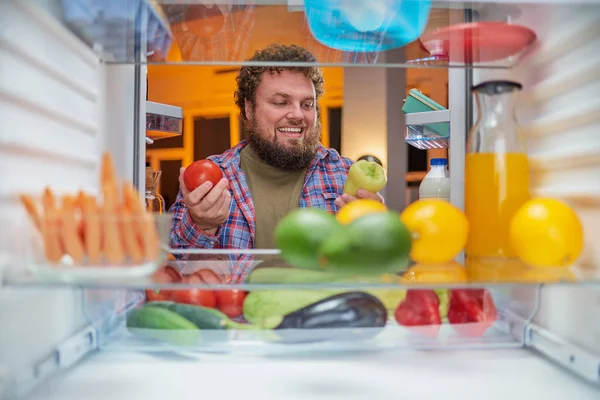 Man choosing what to eat late at night. In one hand paprika and in other tomato. Picture taken from the inside of fridge.