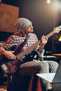 Beyaz delikanlı ev stüdyosunda otururken bas gitar çalmaya kafasına şapka ile.