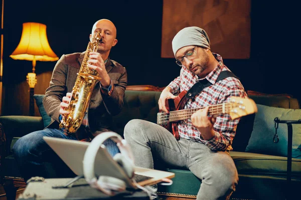 Two talented musicians playing bass gutar and sax while sitting on couch in home studio. — Stock Photo, Image