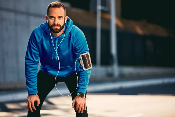 Young Caucasian bearded man standing on a street and resting from running while looking at camera. Sportswear on, earphones in ears. Healthy lifestyle concept.