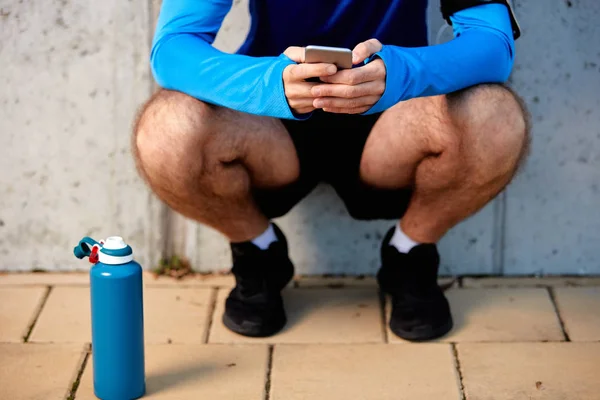Young man crouching and using smart phone. Next to him bottle with water. In background wall. Healthy lifestyle concept.