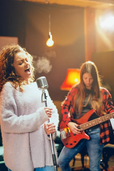 Two women practicing for the gig. Woman with curly hair holding microphone and singing while other one playing bass guitar. Home studio interior.