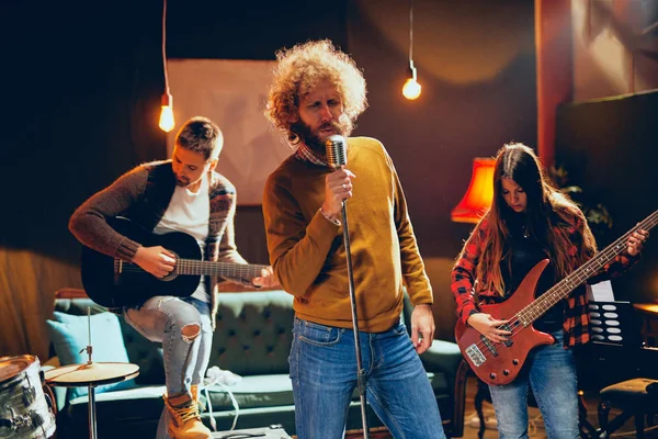 Band practicing for the gig.  Male singer with curly hair holding microphone and singing. In background band playing instruments. Home studio interior.
