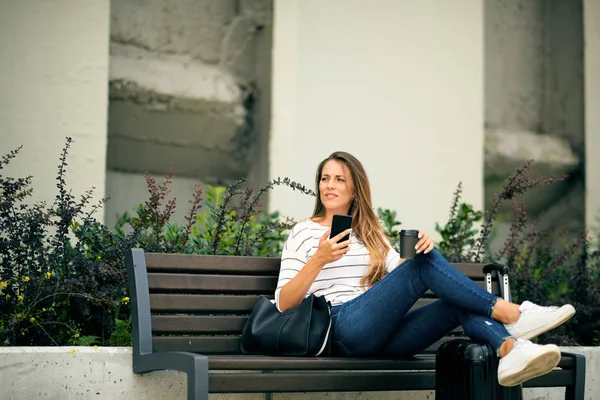 Mujer sentada en el banco y usando un teléfono inteligente mientras espera en la estación. Piernas en el equipaje. En las manos teléfono inteligente y café para llevar . — Foto de Stock