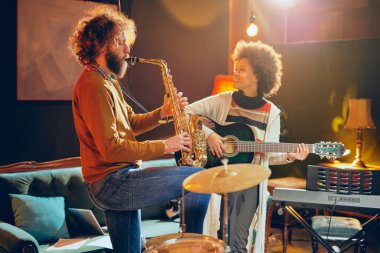 Karışık ırk kadın adam saksafon oynamaya akustik gitar çalmak. Ev stüdyosu iç.