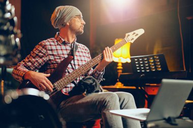 Beyaz delikanlı ev stüdyosunda otururken bas gitar çalmaya kafasına şapka ile.