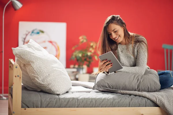 Hermosa morena caucásica sonriente vestida casual sentada en el dormitorio en la cama y usando la tableta . — Foto de Stock