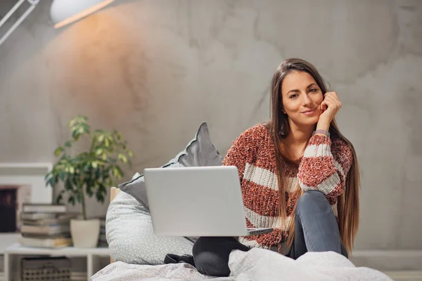 Schöne lächelnde kaukasische Brünette sitzt auf dem Bett im Schlafzimmer, schaut in die Kamera und benutzt Laptop. — Stockfoto