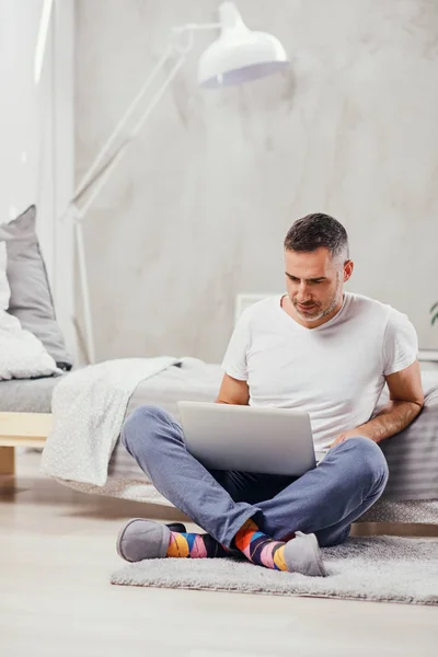 Guapo hombre de mediana edad sentado en el suelo wirh piernas cruzadas, apoyado en la cama y el uso de ordenador portátil . — Foto de Stock