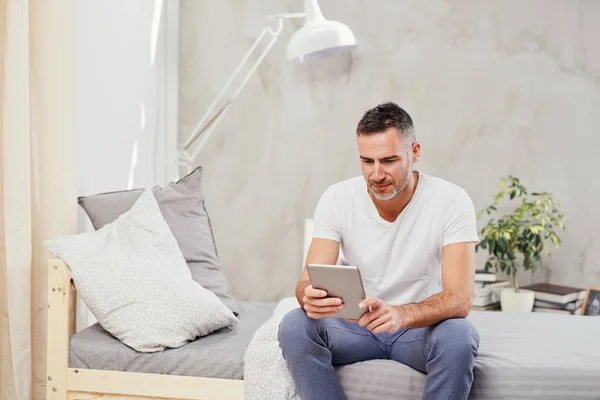 Caucasian man in forties sitting on bed in bedroom and using tablet. — Stock Photo, Image