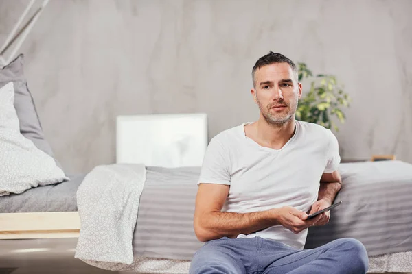 Handsome Caucasian middle-aged man sitting on the floor in bedroom and leaning on bed. In hands tablet. — Stock Photo, Image
