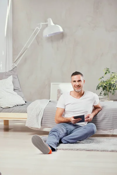 Красивый белый мужчина средних лет, сидящий на полу в спальне и опирающийся на кровать. В руках планшет . — стоковое фото