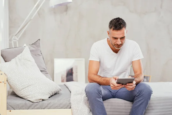 Caucasian man in forties sitting on bed in bedroom and using tablet. — Stock Photo, Image