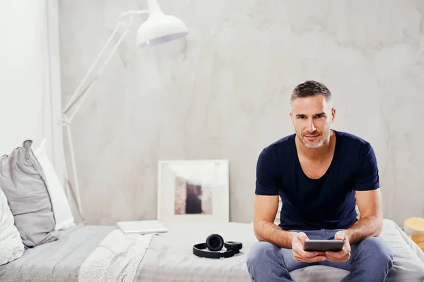 Caucasian man in forties sitting on bed in bedroom and using tablet. — Stock Photo, Image