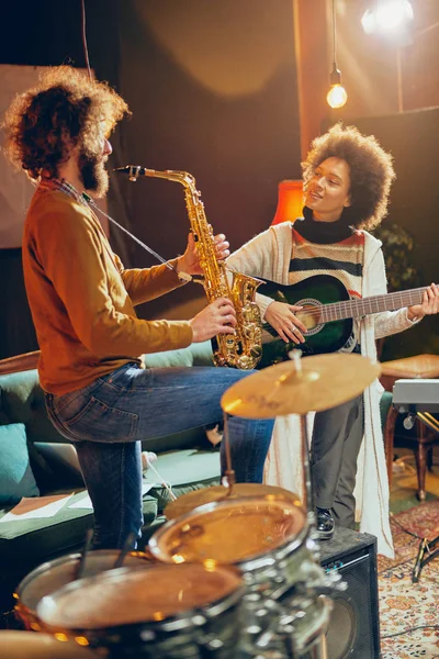 Karışık ırk kadın adam saksafon oynamaya akustik gitar çalmak. Ev stüdyosu iç. — Stok fotoğraf