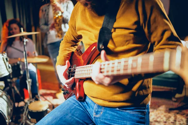 Close up of bass guitarist playing guitar while sitting on the chair. In background rest of the band. Home studio interior. — Stock Photo, Image