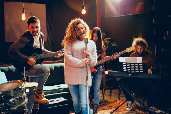 Pratique du groupe pour le spectacle. Femme aux cheveux bouclés tenant microphone et chantant pendant que l'homme en arrière-plan joue de la guitare acoustique. Accueil studio intérieur . — Photo