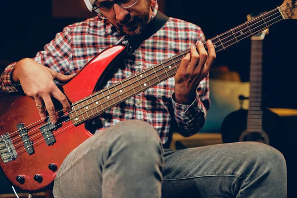 Young Caucasian man with hat on head playing bass guitar while sitting in home studio. — Stock Photo, Image